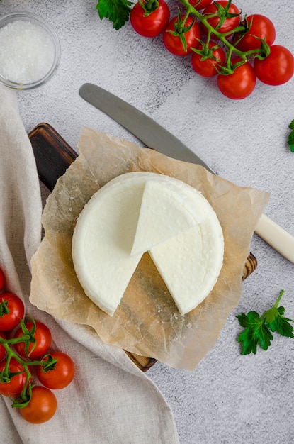 Formaggio a pasta molle o feta fresco fatto in casa su un tagliere di legno con prezzemolo e pomodori ciliegia su un fondo grigio. Orientamento verticale, vista dall'alto