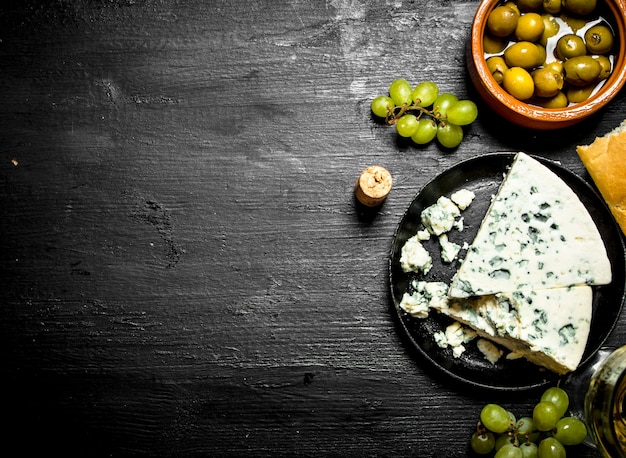 Formaggio blu, olive e uva bianca sulla tavola di legno nera.