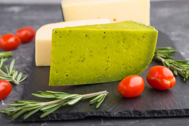 Formaggio verde basilico e vari tipi di formaggio con rosmarino e pomodori su ardesia nera a bordo su una superficie di cemento nera. Vista laterale, da vicino.