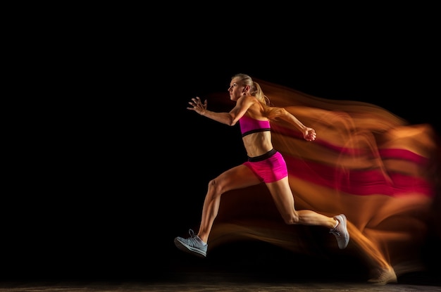 Formazione di corridore di staffetta femminile professionale isolata su sfondo nero per studio in luce mista. Donna in tuta sportiva che si esercita nella corsa. Stile di vita sano, sport, allenamento, movimento, concetto di azione.