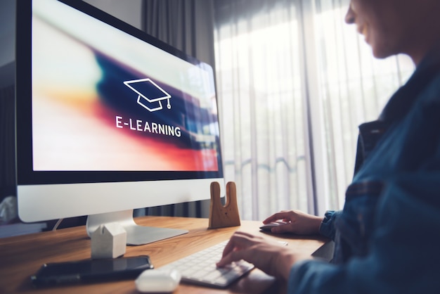 Formazione online, e-learning. La giovane donna è seduta al tavolo, lavorando sul monitor del computer con iscrizione sullo schermo e-learning e l'immagine del berretto accademico quadrato, formazione a distanza.