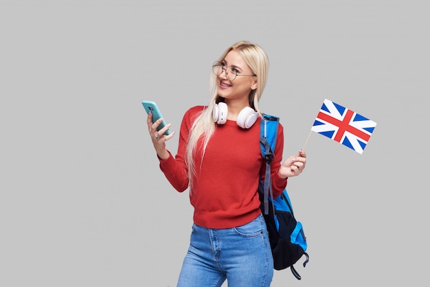 Formazione online, traduttore di lingua straniera, inglese, studente - donna bionda sorridente in cuffia con cellulare e bandiera britannica. Spazio grigio, apprendimento a distanza