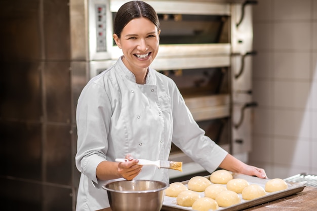 Forno. Donna graziosa sorridente in uniforme bianca che prepara i panini per la cottura in piedi vicino al tavolo
