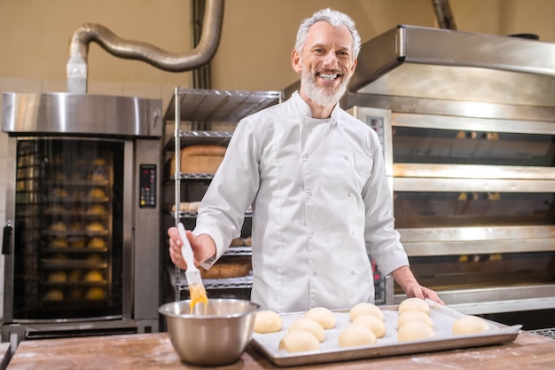 Forno. Uomo barbuto adulto dai capelli grigi sorridente in uniforme bianca che prepara i panini per la cottura in piedi vicino al tavolo