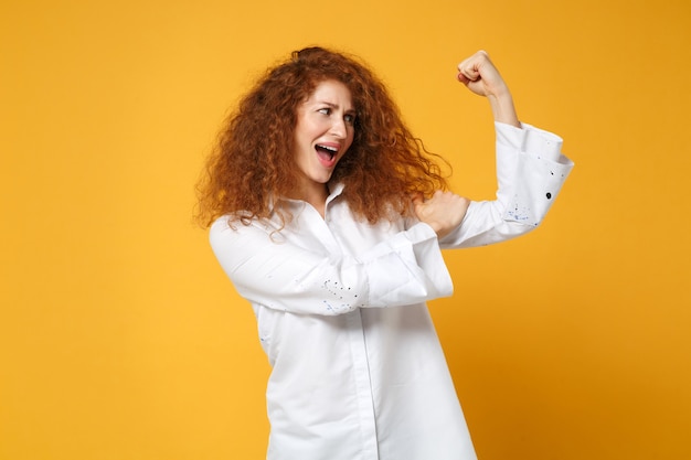 Forte giovane ragazza dai capelli rossi in camicia bianca casual in posa isolata sul muro giallo arancione