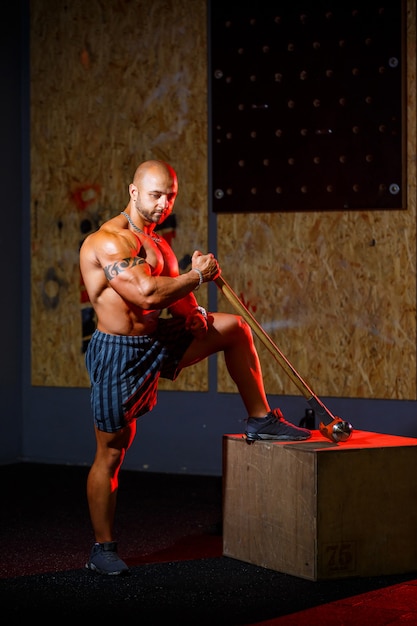 Forte uomo atletico in posa con un martello sportivo sullo sfondo della palestra. Un bodybuilder forte con addominali, spalle, bicipiti, tricipiti e petto perfetti.