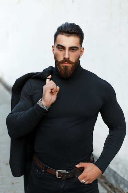 Forte uomo bello in un cappotto nero in una camicia nera con barba e baffi in posa per strada