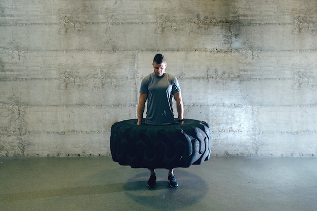 Forte uomo muscoloso in abbigliamento sportivo sollevamento pneumatici mentre in piedi davanti al muro in palestra cross-fit.