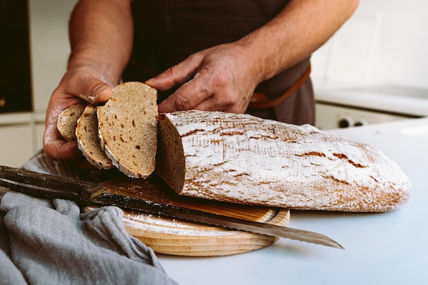 Forti mani muscolari maschili del panettiere, nella farina, tagliano la pagnotta di pane artigianale fatto in casa dalla farina di segale