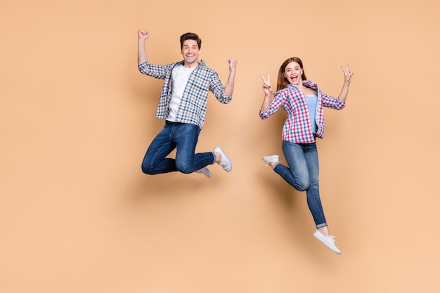 Foto a figura intera di due persone pazza signora ragazzo che salta in alto mostrando v-segno simbolo che celebra successo vincere indossare casual plaid jeans vestiti isolati beige colore di sfondo