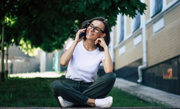 Foto a figura intera di una giovane ragazza, con gli occhiali sul viso, con in mano un telefono, sorridente, che si diverte fuori.