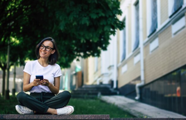 Foto a figura intera di una giovane ragazza, con gli occhiali sul viso, con in mano un telefono, sorridente, che si diverte fuori.
