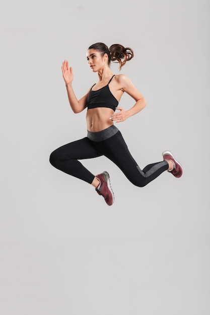 Foto a figura intera nel profilo della donna in buona salute felice in abiti sportivi con gli addominali che saltano e che volano, isolato sopra la parete grigia