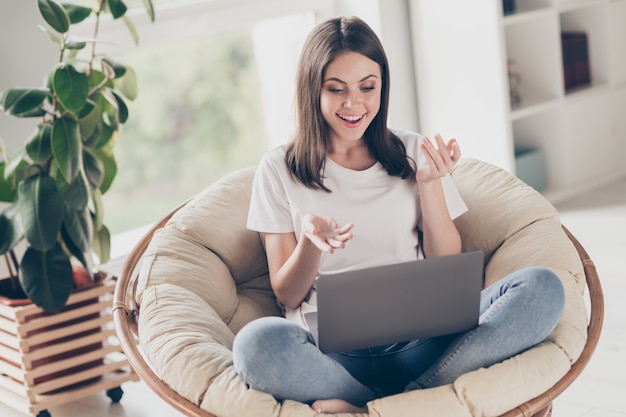 Foto a grandezza naturale di una ragazza positiva che chatta con un video sul laptop, parla, si siede su una sedia di vimini nel soggiorno di casa