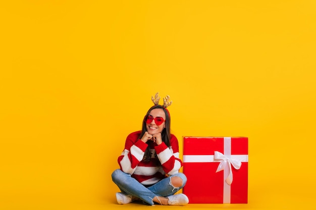 Foto a tutta lunghezza di felice giovane sorridente bella e affascinante donna in corna di renna di Natale mentre è seduta con una grande scatola regalo rossa