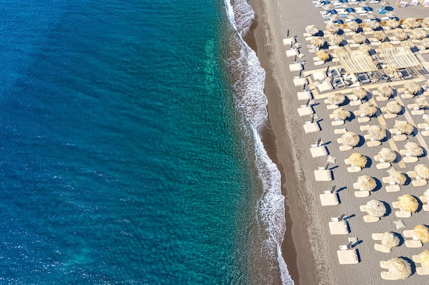 Foto aerea del drone vista dall'alto della spiaggia di Perissa nera con bellissime onde del mare turchese e ombrelloni di paglia Sfondo di viaggio per le vacanze Mar Egeo Isola di Santorini Grecia