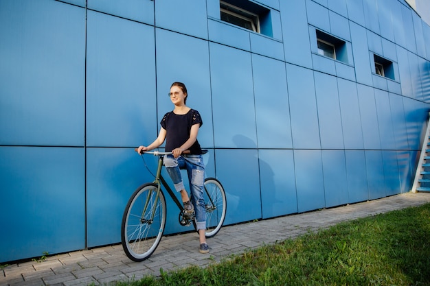 Foto all'aperto della ragazza allegra in occhiali da sole che stanno con la bici contro la costruzione blu. Vestito in eleganti abiti casual.