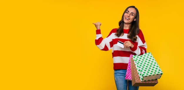 Foto banner di bella giovane donna felice ed eccitata in un maglione a righe con molte borse della spesa colorate e carta di credito nelle mani isolate su sfondo giallo