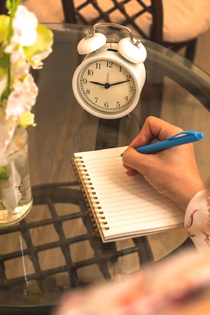 Foto concettuale del diario personale con la donna che scrive sul tavolo di vetro dell'area di lavoro accogliente del taccuino con vista della pianta e della sveglia sopra la spalla