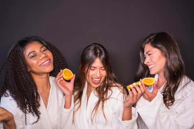 Foto da studio con sfondo grigio di donne felici multietniche in accappatoio che mangiano frutta sana