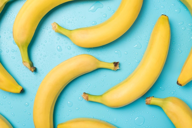 Foto dall'alto di banane gialle non sbucciate e gocce d'acqua su sfondo blu pastello isolato