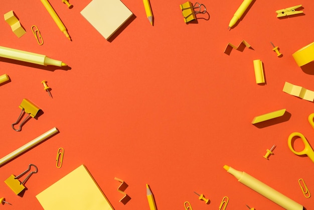 Foto dall'alto di cancelleria gialla forniture scolastiche forbici matite pennarelli fermagli per raccoglitori puntine da disegno e carta adesiva su sfondo arancione vivido isolato con copyspace al centro