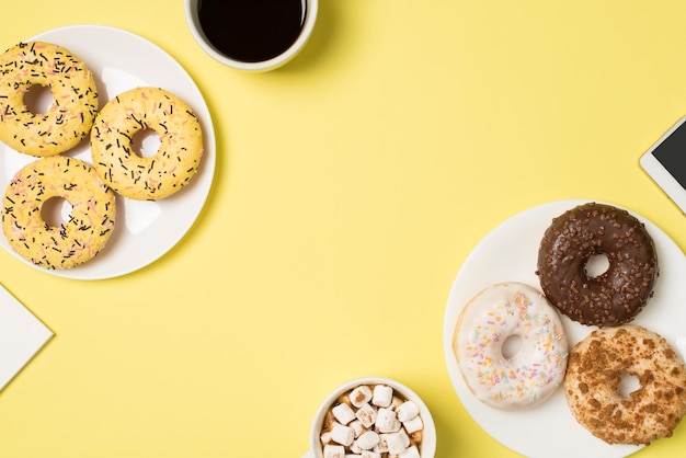 Foto dall'alto di due tazze di bevanda con marshmallow e caffè due piatti con ciambelle di colore diverso e cellulare su sfondo giallo pastello isolato con copyspace