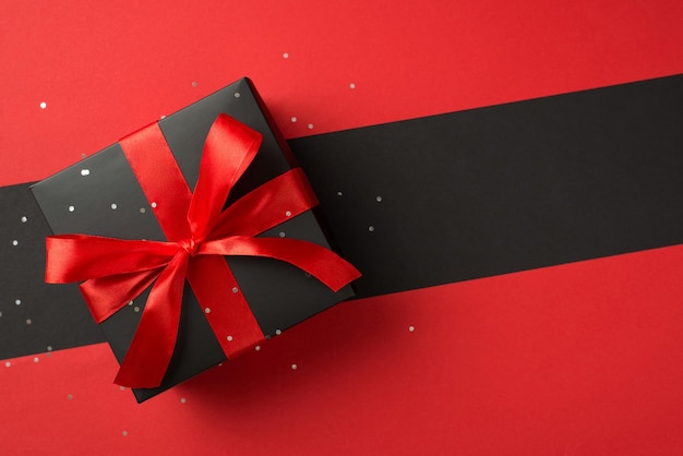 Foto dall'alto di una confezione regalo nera con fiocco di nastro rosso vivo e coriandoli su sfondo bicolore rosso e nero isolato con copyspace