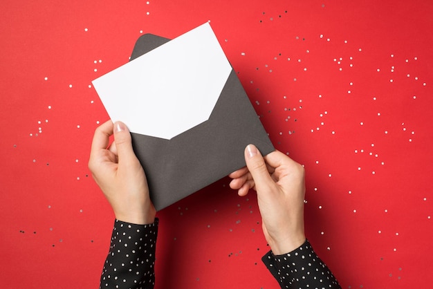 Foto dall'alto in prima persona di mani di donna che tengono una busta nera con una carta bianca all'interno isolata su sfondo rosso con coriandoli