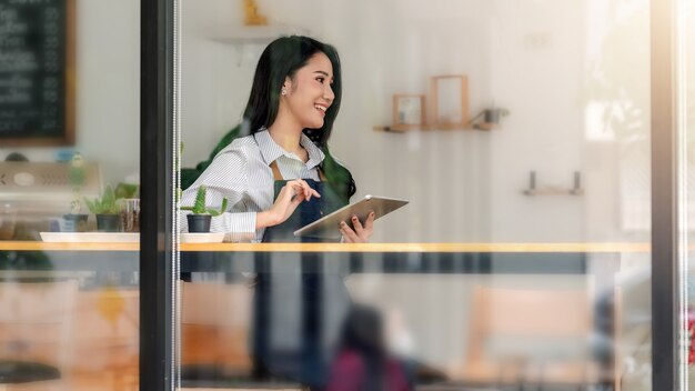 Foto dall'esterno del vetro vista laterale di un piccolo imprenditore una donna asiatica felice seduta al bancone con in mano un tablet in un caffè.