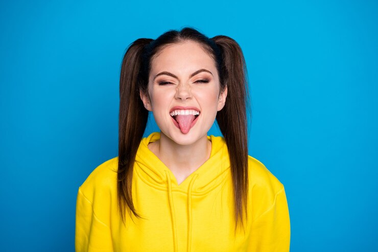 Foto del primo piano di divertente attraente signora divertente due code pettinatura divertente da ragazzina con la lingua fuori bocca umore giocoso indossare casual felpa con cappuccio giallo pullover isolato blu brillante colore di sfondo