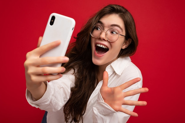 Foto del primo piano di stupefacente bella giovane donna castana positiva che indossa camicia bianca e ottica