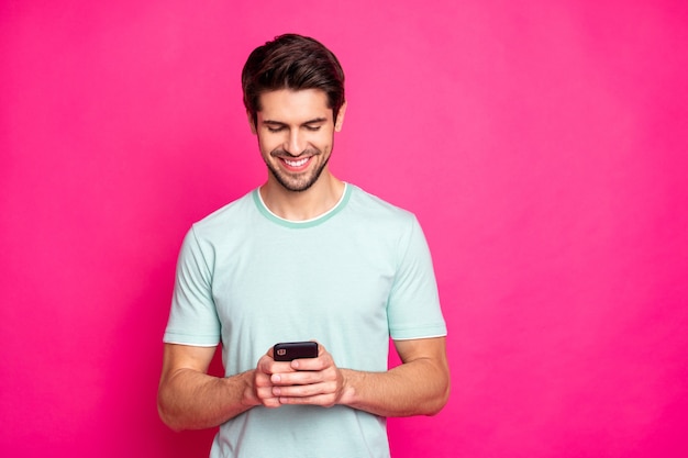 Foto del ragazzo blogger che tiene il telefono nelle mani che controllano gli abbonati sorridente a trentadue denti abbigliamento casual isolato sfondo di colore rosa vivo