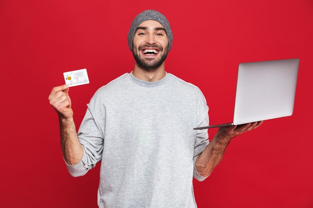 Foto del ragazzo felice anni '30 in abbigliamento casual tenendo la carta di credito e computer portatile d'argento isolato