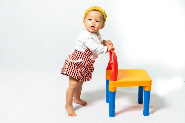 Foto del ritratto di bambino piccolo, 9-10 mesi, ragazza asiatica che indossa camicia bianca con fascia colorata e fascia per capelli gialla, in piedi sulla sedia di plastica colorata, su sfondo bianco.