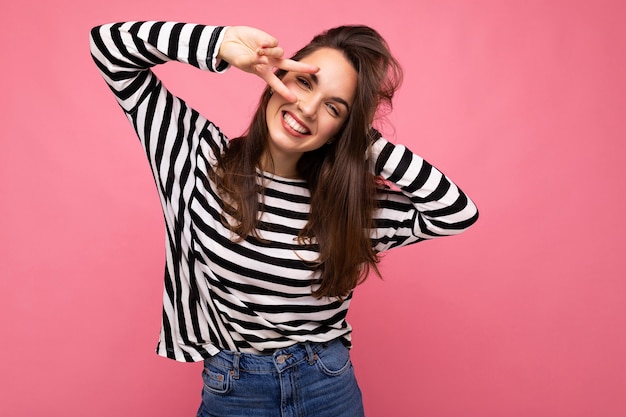 Foto del ritratto di giovane donna castana felice sorridente winsome positiva europea con le emozioni sincere che indossano il pullover a strisce casuale isolato su fondo rosa con lo spazio della copia.