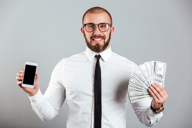 Foto del riuscito uomo 30s in vetri e legame che tiene telefono cellulare e fan delle banconote in dollari, isolato sopra la parete grigia