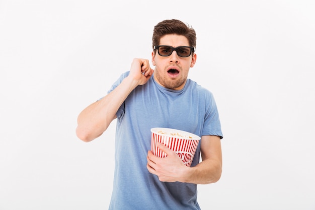 Foto del tipo caucasico in maglietta casuale e vetri 3d che mangiano popcorn e che guardano emozionalmente film nel cinema, isolato sopra la parete bianca