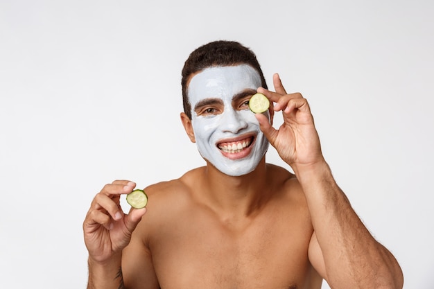 Foto dell'uomo allegro nella maschera cosmetica facciale che prende in giro con il cetriolo isolato sopra bianco