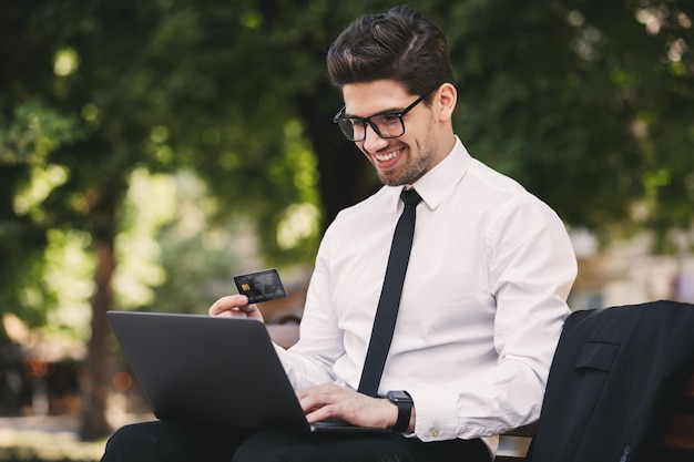 Foto dell'uomo d'affari sorridente in vestito che si siede sulla panchina nel parco verde e lavora al computer portatile durante la giornata di sole