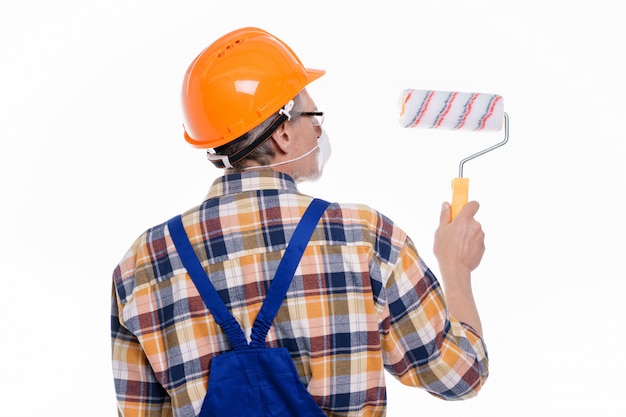 Foto dell'uomo intonacatore posteriore in maschera al lavoro con rullo intonacare il muro degli interni