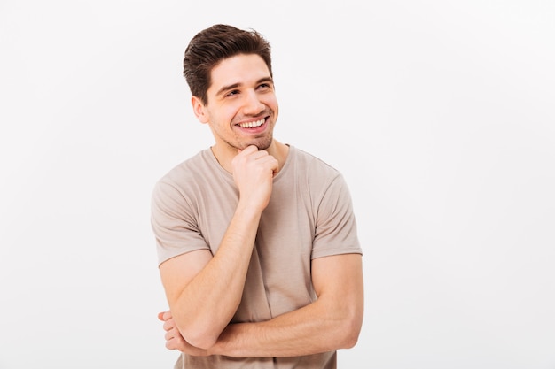 Foto dell'uomo sorridente attraente che sostiene il mento e che osserva da parte, isolata sopra la parete bianca