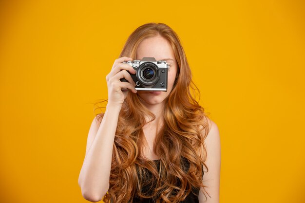 Foto della donna adorabile della testarossa che indossa i vestiti neri che tengono la retro macchina fotografica d'annata e che prendono immagine isolata sopra la parete gialla