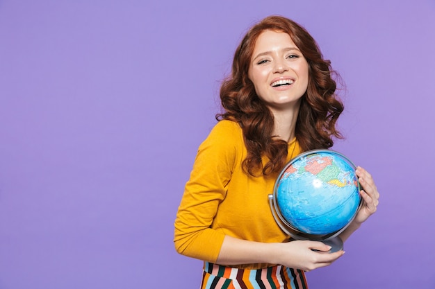 Foto della donna attraente della testarossa che porta i vestiti gialli che sorridono e che tengono il globo della terra sopra la porpora