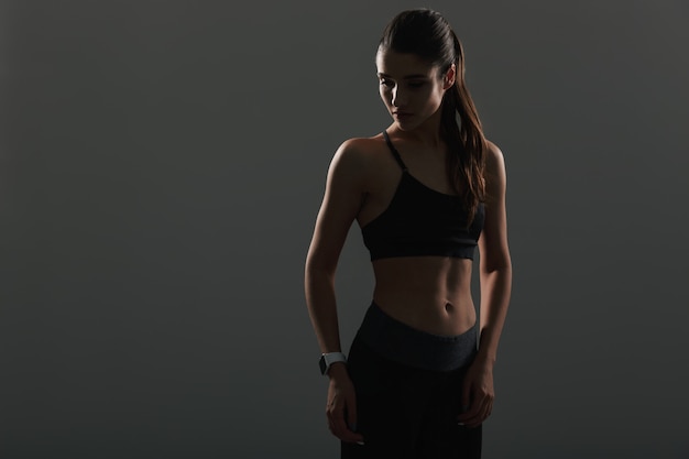Foto della donna graziosa atletica che guarda da parte mentre posando con l'orologio, isolata sopra la parete scura