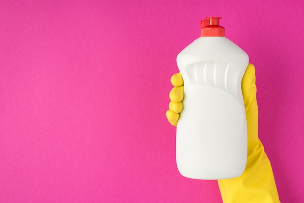 Foto della mano in guanto di gomma gialla che tiene una bottiglia di gel detergente senza etichetta su sfondo rosa isolato con spazio vuoto