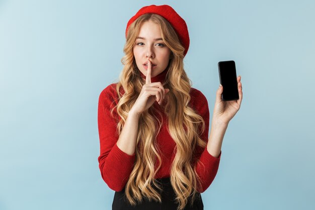 Foto della ragazza timida 20s che indossa il berretto rosso che tiene il telefono cellulare isolato