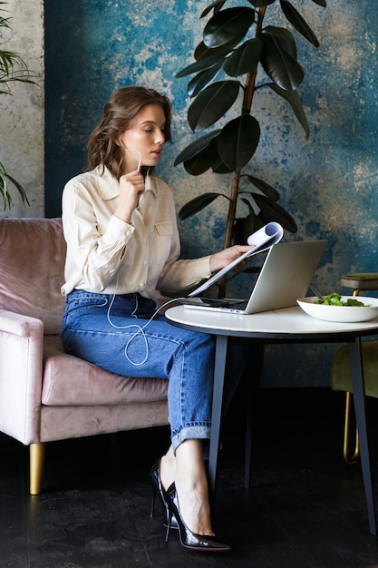 Foto di abbastanza bella giovane donna seduta in un caffè al chiuso utilizzando laptop tenendo i documenti che parlano dal telefono cellulare.