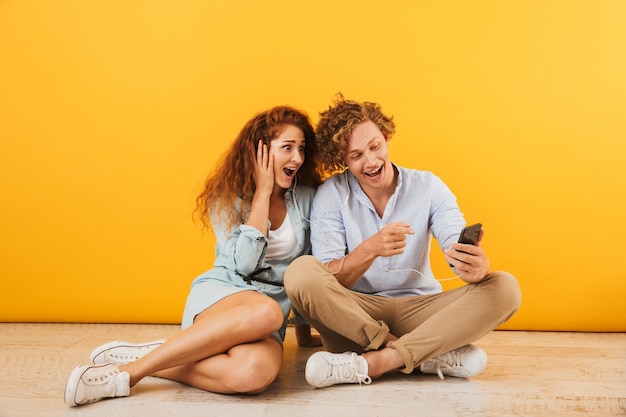 Foto di allegro coppia felice uomo e donna che ascolta la musica tramite gli auricolari mentre è seduto sul pavimento insieme, isolato su sfondo giallo