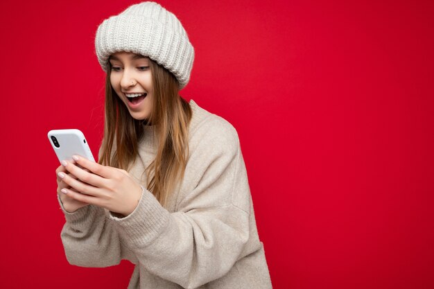 Foto di attraente folle stupito sorpreso giovane donna che indossa abiti casual alla moda in piedi isolato su sfondo con copia spazio tenendo e utilizzando il telefono cellulare guardando lo schermo gadjet.